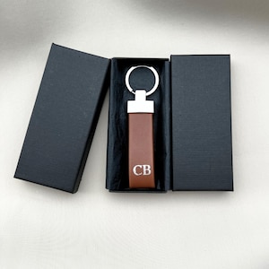 Könnte beinhalten: Ein brauner Leder-Schlüsselanhänger mit silberfarbenen Beschlägen und den Initialen "CB" in Weiß. Der Schlüsselanhänger wird in einer schwarzen Geschenkbox mit offenem Deckel präsentiert. Der Hintergrund ist ein neutrales Off-White.
