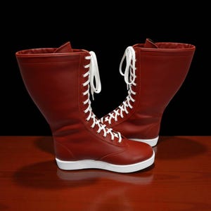 May include: A pair of red leather boots with white laces and soles. The boots have a high-top design and are set against a dark background on a wooden surface.