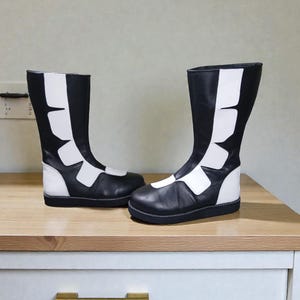 May include: A pair of black and white boots. The boots are knee-high with a flat sole. The design features white panels on the front and sides, creating a striking contrast against the black leather. The boots are displayed on a wooden surface.