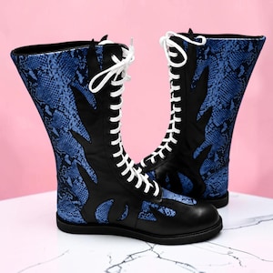 May include: A pair of mid-calf boots featuring a blue and black snakeskin pattern combined with solid black panels. The boots have a front lace-up closure with white laces, a rounded toe, and a flat black sole, extending to the mid-calf height.