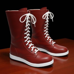 May include: A pair of burgundy-colored boots with white laces and soles. The boots have a mid-calf height and a classic lace-up design. The boots are made of a smooth material and are set against a dark background.