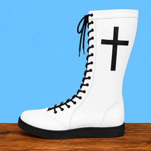 May include: A tall white lace-up boot featuring a black sole and black laces. The side of the boot is decorated with a prominent black cross symbol. The design includes a rounded toe cap and a high shaft that extends well above the ankle area.