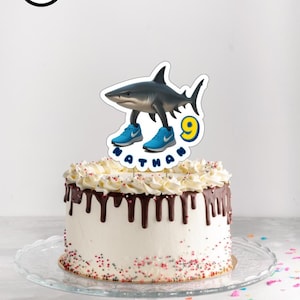 Puede incluir: Una tarta glaseada blanca con goteo de chocolate y chispas de colores. Un adorno para tarta de tiburón con zapatillas azules y el nombre "Nathan" y el número "9" en la parte superior. La tarta está sobre un soporte de cristal. Las palabras "ARCHIVO DIGITAL" están en un círculo negro.