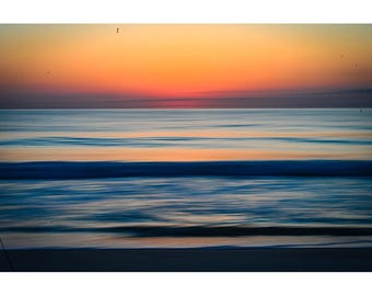 May include: A long exposure photograph of the ocean at sunset. The sky transitions from orange to red, with the ocean waves appearing as blurred blue lines. The beach is visible at the bottom of the frame.
