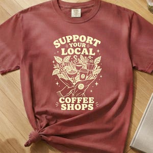 Op de afbeelding: Een chili-kleurig t-shirt met korte mouwen en een crèmekleurige grafische print met de tekst SUPPORT YOUR LOCAL COFFEE SHOPS, vergezeld van illustraties van koffiebonen, een koffiekopje en een mokapot die in twee handen worden gehouden.