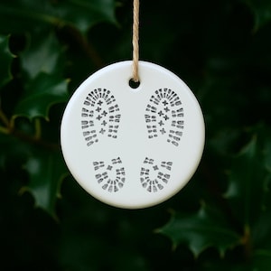May include: A circular white ceramic ornament featuring black boot print designs. It hangs from a natural twine string. The background is a blurred, dark green foliage.