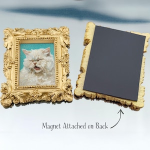 Two ornate rectangular gold frames are shown on a reflective surface. The left frame features a painted portrait of a fluffy white cat with its eyes closed and mouth slightly open, set against a turquoise background. The right frame is flipped to display the back, where a large black magnet is attached, with text below reading “Magnet Attached on Back.