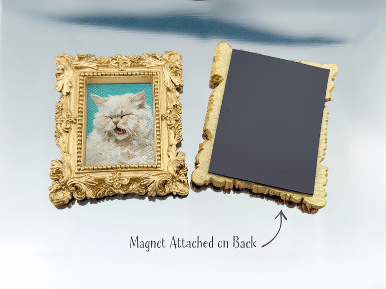 Two ornate rectangular gold frames are shown on a reflective surface. The left frame features a painted portrait of a fluffy white cat with its eyes closed and mouth slightly open, set against a turquoise background. The right frame is flipped to display the back, where a large black magnet is attached, with text below reading “Magnet Attached on Back.