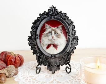 Puede incluir: Retrato enmarcado de un gato con ojos azules y pajarita roja. El marco negro ornamentado, de forma ovalada, se apoya en un soporte decorativo. El fondo es rojo, y el gato es blanco y gris. Decoraciones otoñales en la mesa.