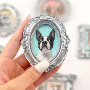Może przedstawiać: Owalna, srebrna ramka z portretem psa rasy Boston Terrier. Pies ma czarno-białe oznaczenia na jasnoniebieskim tle. W tle widoczne są inne oprawione portrety zwierząt.