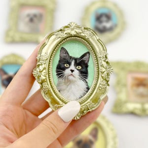 以下が含まれることがあります： 緑色の背景に、白黒の猫が描かれた楕円形の金色の額縁の肖像画。 猫は黄色い目をし、白いマズルをしています。 他の額縁に入ったペットの肖像画が背景にあります。