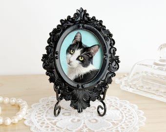 Puede incluir: Un marco de fotos ovalado negro adornado con un retrato de un gato blanco y negro con ojos verdes. El marco está sostenido por un soporte de metal negro y descansa sobre un tapete de encaje blanco. El fondo es azul claro.
