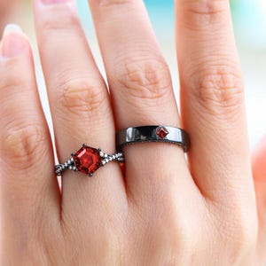 May include: Two black rings with red gemstones. One ring features a large, hexagonal red stone and a band with small white stones. The other ring has a small, square red stone. Jewelry.