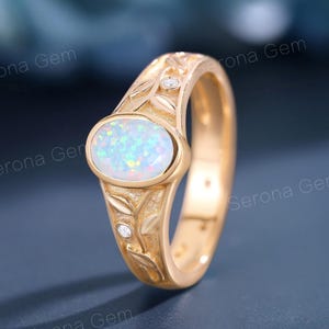May include: A gold ring with an oval opal gemstone and leaf design. The ring has small diamond accents and the text "SERONA GEM". The opal has a white and blue iridescent color.
