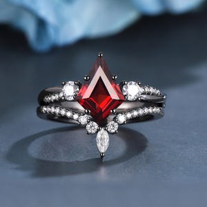 May include: A black engagement ring set featuring a striking red gemstone, kite-shaped, as the centrepiece. The ring is embellished with smaller white gemstones and a delicate band. The set includes a matching band, creating an elegant appearance.