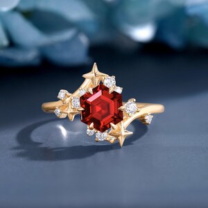 Può includere: Un anello d'oro con una gemma esagonale rosso scuro come elemento centrale. L'anello è adornato con dettagli a forma di stella e piccole gemme trasparenti, creando un design celeste. La fascia dell'anello è in oro lucido.