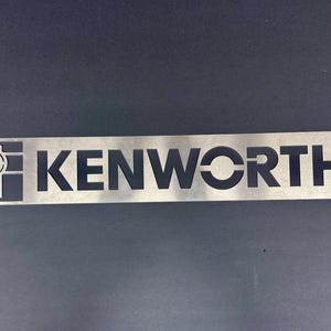 May include: A silver metal sign with a black cutout of the Kenworth logo. The logo features the letters "KENWORTH" and a stylized "K" and "W" inside a circle.