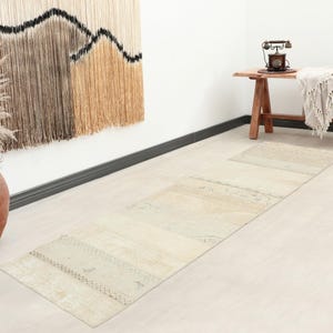 Puede incluir: Una alfombra larga y rectangular con un patrón descolorido en tonos neutros, con sutiles matices beige y crema. La alfombra muestra motivos geométricos y lineales complejos y desgastados en toda su superficie, con una textura envejecida.