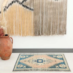 Op de afbeelding: Een rechthoekig tapijt met een geometrisch patroon in blauw, beige en crème. Het tapijt heeft een centraal ruitpatroon en ligt op een lichte vloer. Links staat een grote terracotta vaas met gedroogde planten. Het tapijt is ongeveer 90 cm lang.