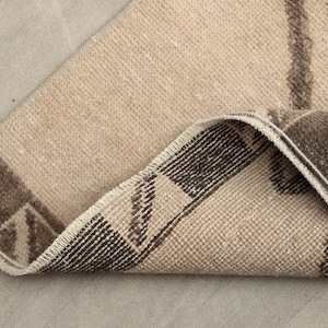 May include: A folded, rectangular rug with a beige and brown color scheme. The rug features a geometric pattern with dark brown borders and accents. The edges are frayed, giving it a vintage appearance. The rug is made of a woven material.