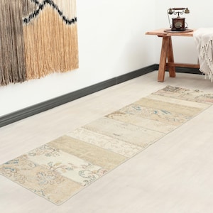 Op de afbeelding: Een lange, rechthoekige loper met een patchworkontwerp in gedempte beige, crème en vervaagde blauwe tinten met bloemmotieven. Het kleed bestaat uit meerdere aan elkaar genaaide secties, elk met ingewikkelde, verweerd ogende decoratieve patronen.