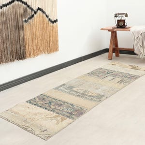 Op de afbeelding: Een rechthoekige patchwork loper met een beige, crème en gedempte blauwe kleurstelling. De loper is gemaakt van verschillende rechthoekige stukken die aan elkaar zijn genaaid. De loper ligt op een lichtgekleurde houten vloer. De loper is ongeveer 244 cm lang.