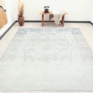 May include: A large rectangular area rug featuring a faded, light blue and grey distressed pattern with a central medallion and an intricate border design. The rug has a low-pile texture and covers the floor, displaying a worn, vintage-inspired aesthetic.
