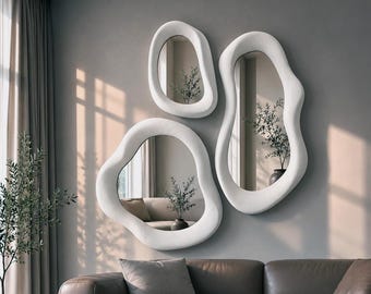 May include: A set of three wall mirrors with irregular, organic, white frames. The mirrors are mounted on a wall above a brown leather sofa, reflecting a room with a plant and natural light. The frames have a smooth, rounded, and asymmetrical shape.