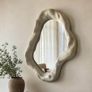May include: An irregularly shaped, off-white wall mirror with a textured, organic frame. The mirror hangs on a neutral-colored wall, reflecting a room with a chair and curtains. A wooden cabinet with decorative items sits below the mirror.