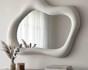 May include: A wall-mounted mirror with an irregular, organic, wavy white frame. The mirror reflects a window with sheer curtains. The frame has a smooth, matte finish and a thick, rounded profile that creates a fluid, asymmetrical shape.