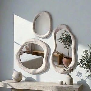 Pode incluir: Um conjunto de três espelhos de parede com molduras de formas orgânicas irregulares e acabamento texturizado em branco sujo. Os espelhos estão dispostos em grupo, refletindo o interior de um quarto com uma cama e uma planta, sobre uma mesa de console de pedra.