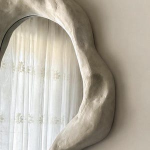 May include: A large, irregularly shaped mirror with a light gray, textured frame. The mirror reflects a sheer white curtain with a floral pattern. The frame has a unique, organic design, adding a modern touch to the decor.