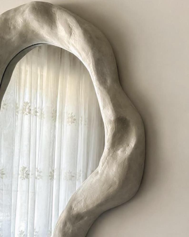 May include: A large, irregularly shaped mirror with a light gray, textured frame. The mirror reflects a sheer white curtain with a floral pattern. The frame has a unique, organic design, adding a modern touch to the decor.