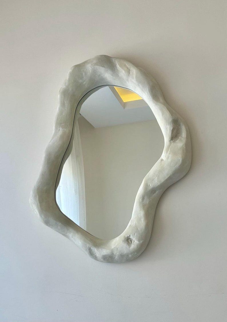 May include: An organically shaped wall mirror with a textured, off-white frame. The mirror's irregular form adds a modern, artistic touch to interior decor. The frame appears to be made of a plaster-like material, creating a unique aesthetic.