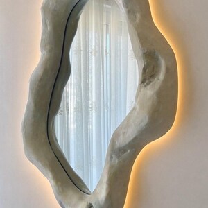 May include: An organically shaped wall mirror with a textured, light gray frame. The mirror has a warm, glowing light around the edges, creating a soft ambiance. The design is modern and artistic, suitable for home decor.