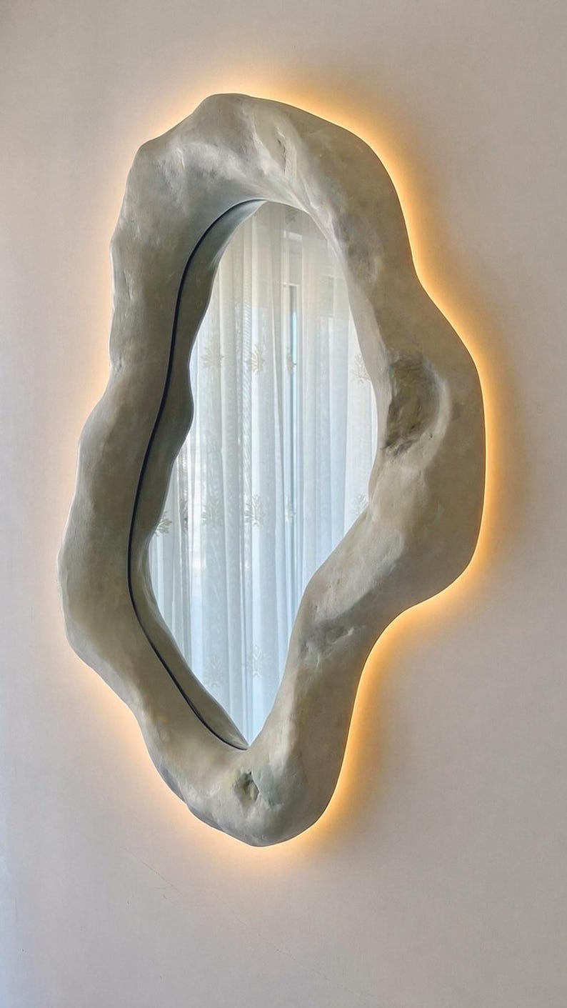 May include: An organically shaped wall mirror with a textured, light gray frame. The mirror has a warm, glowing light around the edges, creating a soft ambiance. The design is modern and artistic, suitable for home decor.