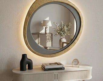 May include: An oval wall mirror with an irregular, textured gold frame and integrated LED backlighting. The mirror features a smooth gray inner border and is mounted above a light-colored, fluted console table with four slender legs.