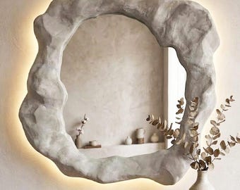 May include: A wall-mounted mirror featuring an irregular, organic-shaped frame with a textured, light gray stone-like finish. The mirror includes integrated backlighting that casts a soft glow around the perimeter of the sculpted frame.
