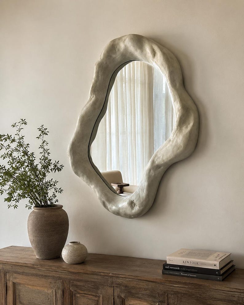 May include: An irregularly shaped, off-white wall mirror with a textured, organic frame. The mirror hangs on a neutral-colored wall, reflecting a room with a chair and curtains. A wooden cabinet with decorative items sits below the mirror.