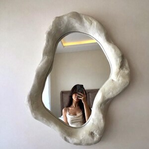 May include: An irregularly shaped mirror with a textured, off-white frame. The mirror reflects a person taking a selfie. The frame has a unique, organic design, adding a modern touch to the wall decor.