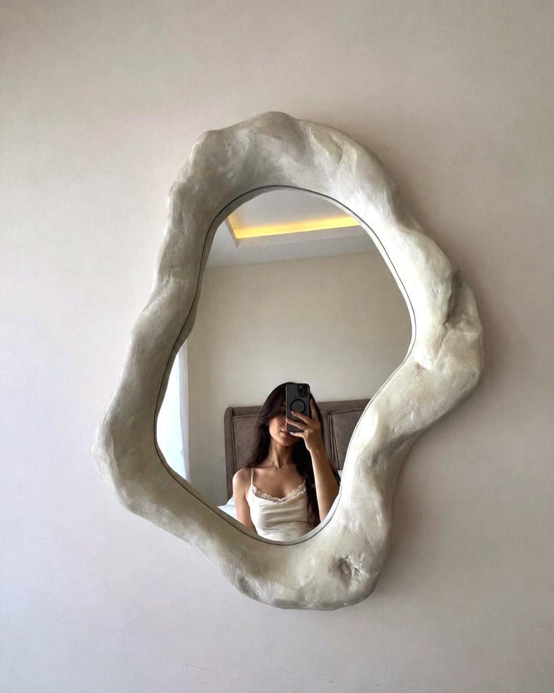 May include: An irregularly shaped mirror with a textured, off-white frame. The mirror reflects a person taking a selfie. The frame has a unique, organic design, adding a modern touch to the wall decor.