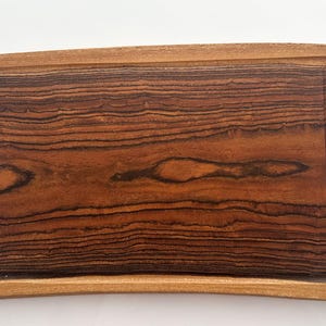 May include: A rectangular wooden chopping board with a curved edge. The board has a dark brown wood grain pattern with lighter brown accents. A recessed groove runs along the top edge.