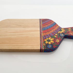 May include: Wooden serving board with a natural wood finish on the left and a colourful, hand-painted design on the right. The design includes flowers and geometric patterns in red, yellow, blue, and purple. The board has a handle.