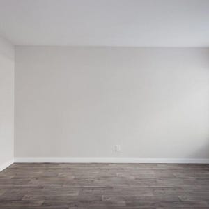 Pode incluir: Um quarto vazio com paredes cinza claro e um chão escuro com aspecto de madeira. Uma janela com persianas brancas está no lado direito. O quarto é bem iluminado, sugerindo luz natural.