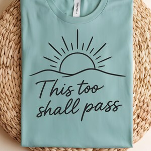 May include: A light teal t-shirt with a black sun and wave design, and the words "This too shall pass" in a script font. The shirt is folded on a woven, light brown surface. The words "Summer Vibes" are in the upper right corner.