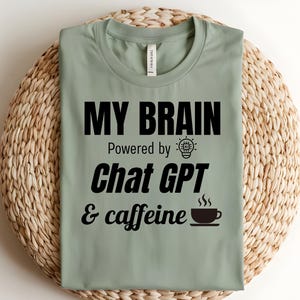 Op de afbeelding: Een saliegroen T-shirt met zwarte tekst die "My Brain Powered by Chat GPT & caffeine" luidt, met een gloeilamp en een kopje koffie-icoon.