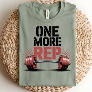 May include: A sage green t-shirt with the text "ONE MORE REP" in black and red. Below the text is a barbell with red weights. The shirt is displayed on a woven surface.