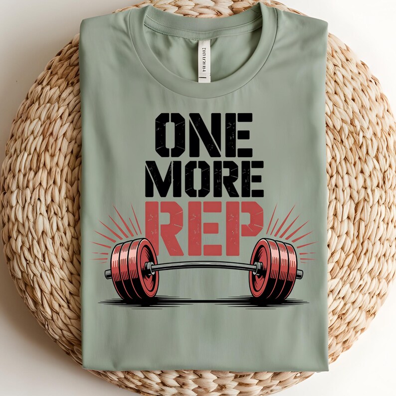 May include: A sage green t-shirt with the text "ONE MORE REP" in black and red. Below the text is a barbell with red weights. The shirt is displayed on a woven surface.