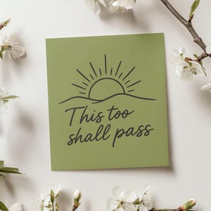 May include: A square, olive-green card with the phrase "This too shall pass" in a handwritten font. The card features a black line drawing of a sun rising over a horizon. White flowers and branches surround the card.