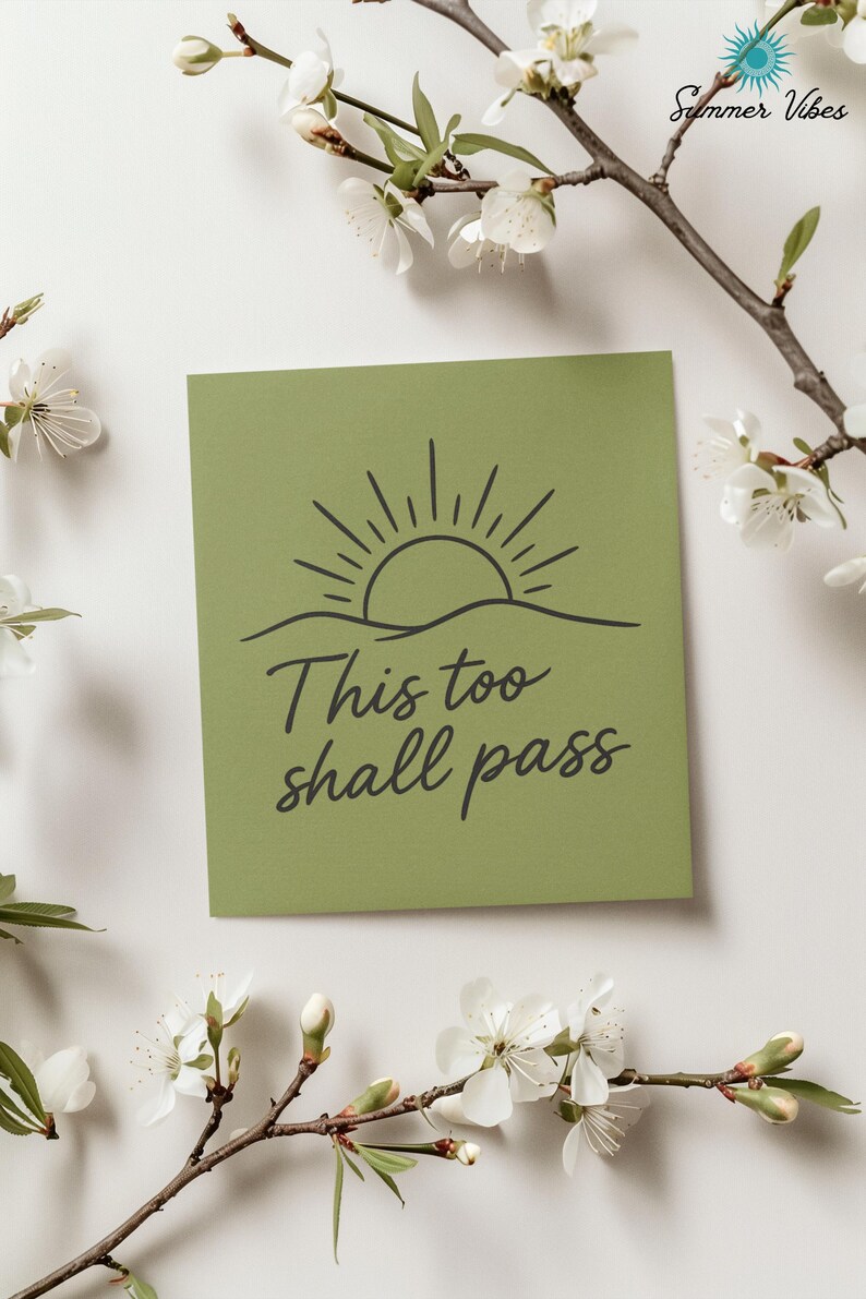 May include: A square, olive-green card with the phrase "This too shall pass" in a handwritten font. The card features a black line drawing of a sun rising over a horizon. White flowers and branches surround the card.